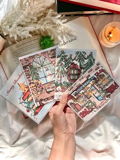 a person is holding some christmas cards in their hand while sitting on a bed next to a candle