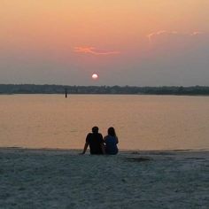 two people are sitting on the beach watching the sun go down over the water at sunset