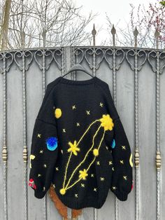 a black sweater with yellow stars and planets on it hanging from a metal fence next to a tree