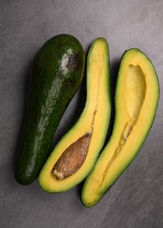 two avocados cut in half on a gray surface with one sliced open and the other whole