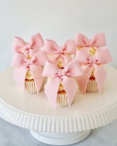 four cupcakes with pink bows are on a white cake plate