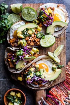 three tacos on a cutting board with avocado and limes next to them