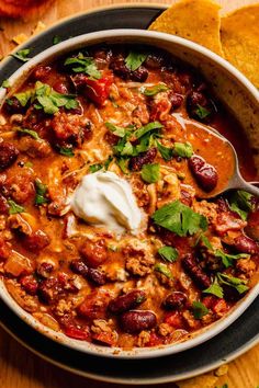 a bowl filled with chili and sour cream next to tortilla chips on a wooden table