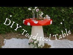 a red and white table with flowers on it, sitting in front of some bushes