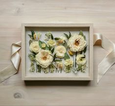 an arrangement of flowers is displayed in a shadow box on a wooden table with ribbon