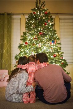 three people sitting in front of a christmas tree with their arms around each other's shoulders