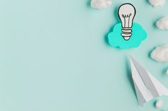 a paper airplane next to a light bulb and clouds on a blue background with copy space