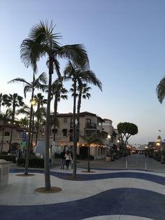 palm trees are in the foreground and people walk on the sidewalk near buildings at dusk