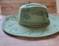 This hat is sold out, but we can create a similar one.. This ones got a "Coastal Cowgirl" Vibe.   "Happiness Comes In Waves"  This one of a kind Fedora is decorated with Palm Trees, Waves, Mermaid Tail and a sand dollar.  This is perfect for the #BarefootCountryMusicFest.  Concert & Rodeo season is just around the corner.    She is screaming Vacation.  This is the only one I have in this color, but we can do this design on other colors. Country Style Wide Brim Felt Hat For Beach, Summer Country Style Felt Hat For Beach, Summer Beach Country Style Felt Hat, Western Style Felt Hat For Beach In Summer, Western Brimmed Felt Hat For Beach, Western Style Brimmed Felt Hat For Beach, Custom Summer Felt Hat For Beach, Summer Felt Cap, Western Felt Hat For Vacation