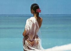 a woman with a flower in her hair standing on the beach looking out at the ocean