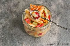 a glass jar filled with pasta salad on top of a stone counter next to a spoon