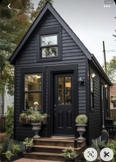 the tiny house is painted black and has two plants in pots on the front porch