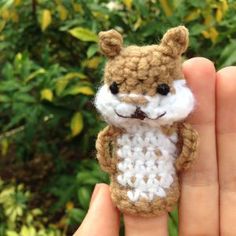 a small crocheted teddy bear sitting on top of someone's hand in front of some bushes