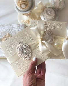 a hand holding two cards with bows and brooches on them next to lace doily