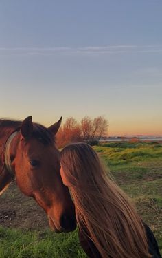 a woman standing next to a brown horse on top of a lush green field under a blue sky