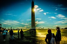 people are walking around in front of the washington monument at sunset, with bright sun peeking through clouds