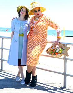 two women standing next to each other on a pier near the ocean wearing hats and dresses