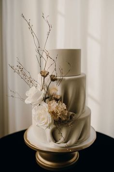 a three tiered cake with flowers on top is sitting on a gold pedestal in front of a window