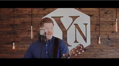 a man holding a guitar and singing into a microphone in front of a wooden wall