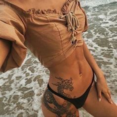 a woman with tattoos on her stomach standing in front of the water at the beach