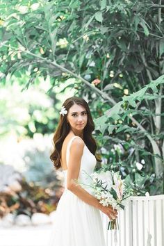 a woman standing next to a white fence holding a bouquet in her hand and looking at the camera