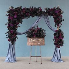 a wooden sign sitting under a purple flower covered arch with draping on it