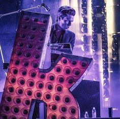 a man standing in front of a giant letter on stage with lights behind him and sound equipment around his neck