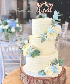 a white wedding cake with blue flowers and greenery sits on a tree stump at the reception table