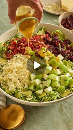 a person pouring dressing into a large bowl filled with vegetables and fruit, along with other food items