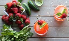 two cocktails with strawberries and cucumber garnish sit on a wooden table