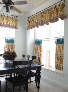 a dining room table and chairs in front of two windows with valances on them