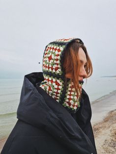 a woman with red hair wearing a crocheted hood on the beach looking at the water