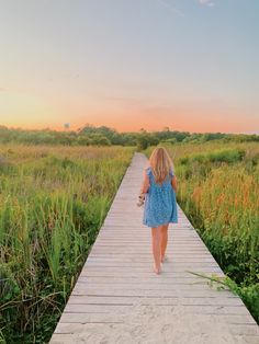 a woman walking down a wooden walkway in the middle of a grassy field at sunset