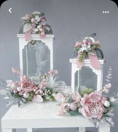 two white lanterns with pink flowers and greenery on the top are sitting on a table