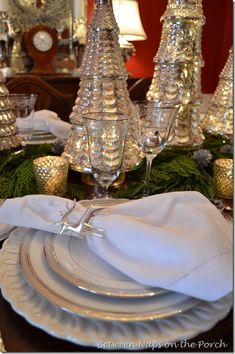 a dining room table set for christmas with silver and white dishes, candlesticks and pine cones