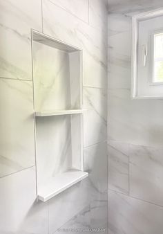 a bathroom with white marble walls and shelves on the wall, next to a window