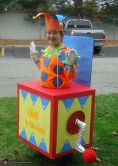 a child in a clown costume sitting on top of a cube shaped box with his hands up