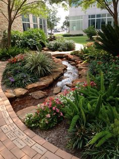 a garden with flowers and plants around it, along with a brick walkway that leads to the front door