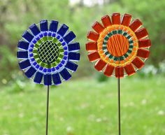 two colorful glass flowers on top of wooden sticks in the grass with trees in the background