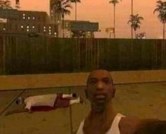 a man in white shirt standing next to a skateboard on cement area with fence and palm trees