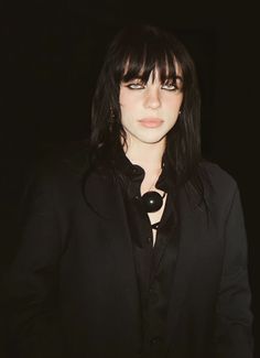 a woman with long black hair and bangs wearing a black suit, standing in the dark