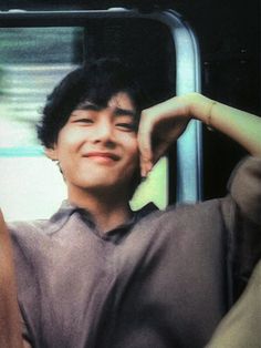 a young man sitting on a train holding his hand up to his head and smiling