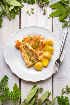 a white plate topped with fish and lemons next to green leaves on top of a wooden table