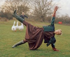 a woman in medieval garb is doing a handstand on the grass with geese behind her