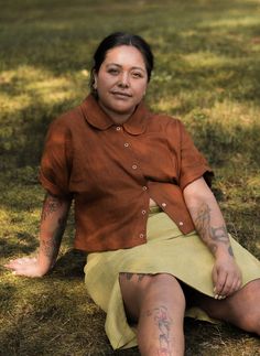 a woman sitting in the grass with tattoos on her arm and leg, wearing a skirt