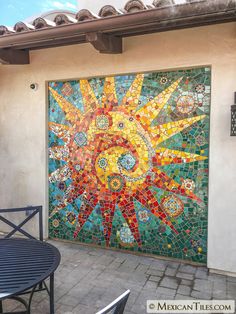 a mosaic tile mural on the side of a building with chairs and table in front of it