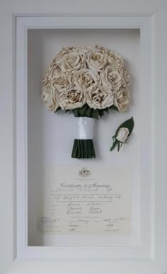 a bouquet of white roses is in a frame with a certificate on the wall behind it