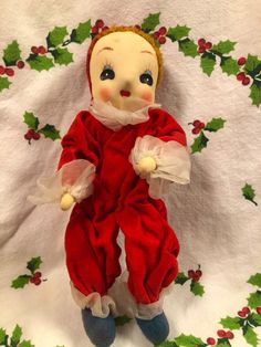 an old fashioned christmas doll is sitting on a white sheet with holly wreaths around it