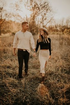 a man and woman holding hands walking through tall grass in an open field at sunset