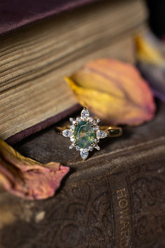 The "Azalea" engagement ring from Staghead Designs sits on a stack of vintage books. This 14K yellow gold engagement ring features an oval cut moss agate center stone & starburst diamond halo with marquise & round diamond accents. Whiskey Barrel Wedding Ring, Antler Wedding Rings, Agate Wedding Ring, Wood Wedding Ring, Moss Agate Engagement Ring, Agate Engagement Ring, Mens Engagement, Colored Stone, Ring With Diamond